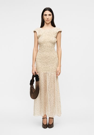 Γυναίκα με μακριά σκούρα μαλλιά που φορά ένα μπεζ φόρεμα με υφή και διαφανές πουά φούστα, κρατώντας καφέ τσάντα και φορώντας καφέ ψηλοτάκουνα.