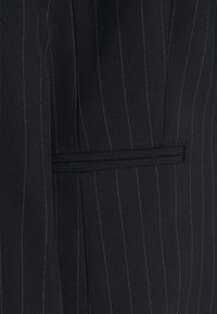 Ciemna tkanina z cienkimi, pionowymi białymi prążkami oraz poziomą kieszenią z patką na dopasowanym ubraniu.