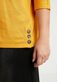 Camiseta de algodón amarilla con dobladillo redondeado, que presenta tres acentos de botones grises en el lado. La tela tiene un acabado texturizado.