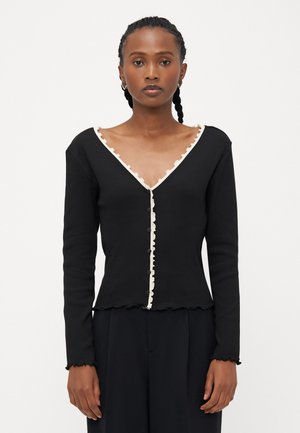 Svart ribbad cardigan med långa ärmar, V-ringning och krämfärgad flikig dekor. Har en knäppning framtill och en beskuren nederkant.