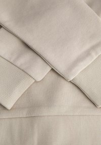 Dettaglio di una felpa color beige caratterizzata da una texture liscia, polsini a coste e una fascia orizzontale a coste in vita, sovrapposta per un effetto di profondità visiva.