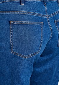 Jeans en denim bleu foncé avec une poche arrière au sommet courbé, cousue avec un fil orange contrastant. Texture douce, design classique à cinq poches.