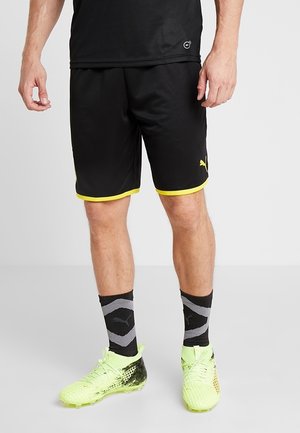 Hombre vestido con pantalones cortos deportivos negros con ribetes amarillos, calcetas negras de Puma y zapatillas neón verdes, de pie contra un fondo liso.