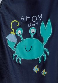 Applique de crabe turquoise sur un tissu foncé, avec des détails brodés. Présente un visage souriant, un hameçon et de petits motifs de poissons.