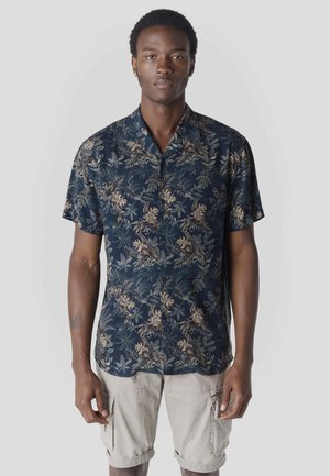 Hombre con camisa de manga corta azul marino con patrón floral beige y pantalones cortos cargo beige claro, de pie contra un fondo liso.