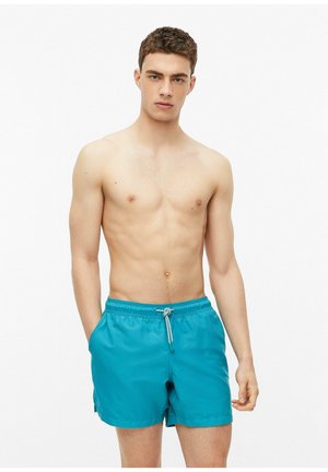 Jeune homme aux cheveux courts et bouclés portant un short de bain turquoise avec cordon, debout sur fond blanc les mains dans les poches.