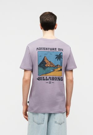 Άτομο που φοράει λιλά μπλούζα με γραφικά βουνού, φοινικόδεντρου και παραλίας και το κείμενο "Adventure Div. Billabong" στο πίσω μέρος.