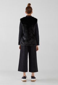 Kvinna med håret i en knut, iförd en svart fuskpälsväst, svarta vida cropped byxor och svart-vita sneakers, stående vänd bort.