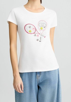 Kobieta w białej koszulce z krótkim rękawem z różową grafiką rakiety i piłki tenisowej, połączona z luźnymi jasnoniebieskimi dżinsami.