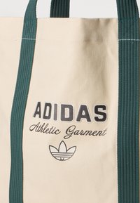Bežs iepirkumu soma ar tumši zaļām rokturiem, uz kuras ir rakstīts "ADIDAS Athletic Garment" un redzams Adidas trefoil logo melnbaltā krāsā.