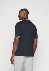 Polo nero con colletto e polsini a righe bianche, realizzato in un tessuto liscio. Indossato con pantaloni della tuta grigi chiari.