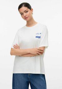 Biała koszulka z krótkim rękawem z niebieskim nadrukiem tekstowym i wzorem na lewej piersi, wykonana z bawełny o luźnym kroju.