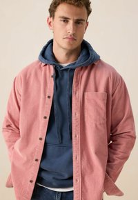 Rode corduroy shirt met een kraag, voorzak en drukknopen over een blauwe hoodie met trekkoord. Draagt het met donkere spijkerbroek.