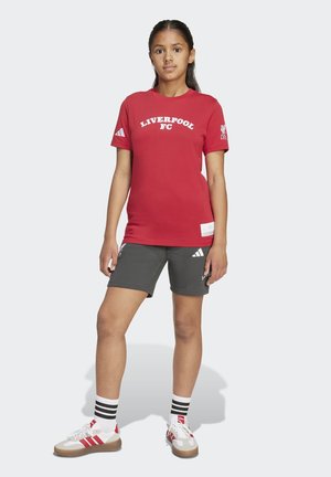 Flicka som bär en röd Liverpool FC t-shirt, grå sportshorts, vita Adidas-sneakers och vita strumpor med svarta ränder, stående mot vit bakgrund.