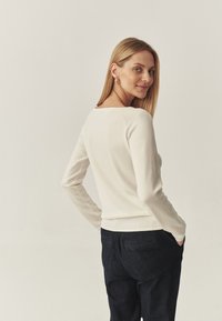 Pulover tricotat de un bej deschis, cu mâneci lungi și decolteu în formă de semicerc, asortat cu pantaloni din denim de culoare albastră închis, cu croi clasic și buzunare la spate.