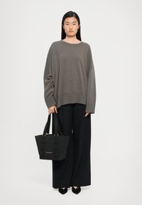 Žena u labavom taupe džemperu i crnim širokim hlačama, drži crnu Love Moschino torbu, naslonjena na jednostavnu svijetlu pozadinu.