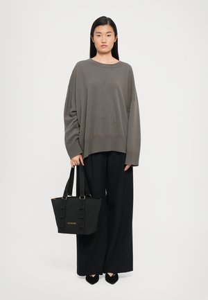 Vrouw in een losse taupekleurige trui en zwarte wijde broek, met een zwarte Love Moschino tote bag, tegen een effen lichte achtergrond.