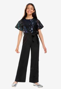 Svart vida byxdress med en paljettkortärmad topp i iriserande färger och en rosettdetalj vid midjan. Silverfärgade ballerina-skor fulländar looken.