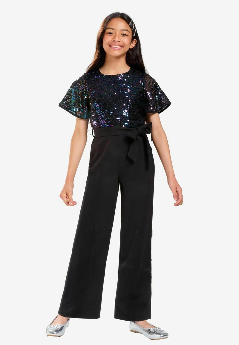 Svart vida byxdress med en paljettkortärmad topp i iriserande färger och en rosettdetalj vid midjan. Silverfärgade ballerina-skor fulländar looken.