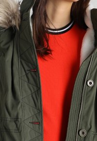 Parka verde oliva con capucha de piel sintética, con detalles acolchados y herrajes metálicos. Debajo lleva una camiseta roja de canalé con un escote en negro y blanco.