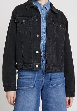 Persoon draagt een zwart spijkerjack over een blauwe spijkerblouse met knopen en een lichtblauwe, hoogwater spijkerbroek tegen een effen grijze achtergrond.