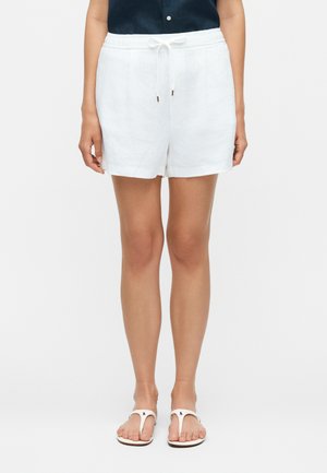 Γυναίκα που φοράει λευκά σορτς με κορδόνι, μπλε μπλούζα μέσα από το παντελόνι και λευκά σανδάλια, στέκεται σε απλό ανοιχτό φόντο.