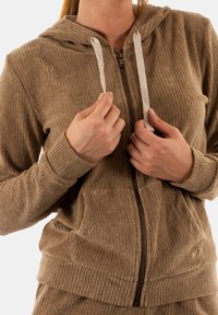 Sudadera con cremallera de color marrón ribeteada y con un tejido texturizado, dos bolsillos frontales y cordones en contraste; cuenta con capucha y cierre de cremallera.