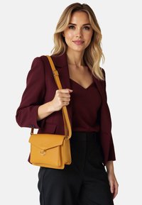 Borsa a tracolla in pelle gialla senape con pattina frontale, dettaglio anello in metallo, finitura testurizzata e tracolla regolabile, indossata con un blazer scuro.