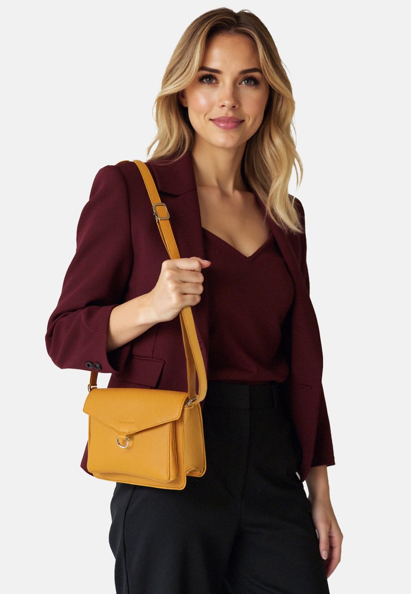 Borsa a tracolla in pelle gialla senape con pattina frontale, dettaglio anello in metallo, finitura testurizzata e tracolla regolabile, indossata con un blazer scuro.