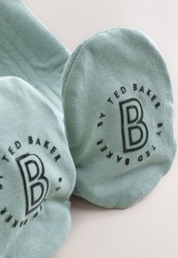 Turkis sokker lavet af blødt stof, med afrundet tå og præget logo-design med teksten "TED BAKER" og et fremtrædende "B" i midten.