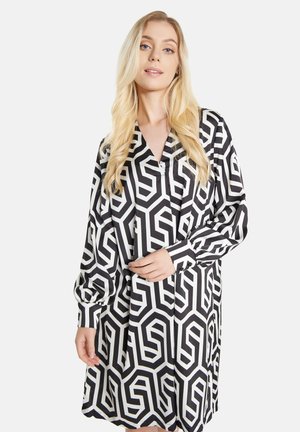 Femme blonde portant une robe à manches longues à motifs géométriques noirs et blancs avec un col en V, debout devant un fond blanc uni.