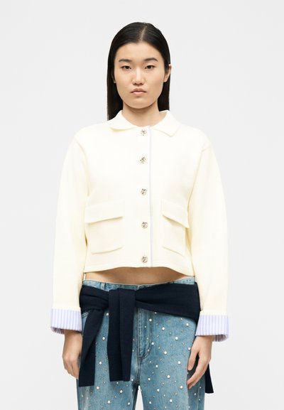 Jeune femme portant une veste courte crème avec des boutons dorés, des poignets rayés, un jean orné de perles, et un pull sombre noué autour de la taille.