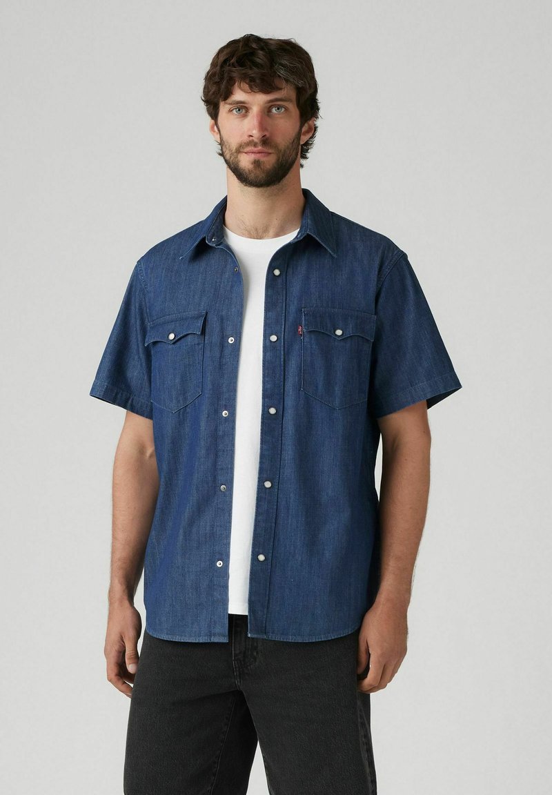 Camisa de mezclilla de manga corta en azul oscuro con dos bolsillos frontales, botones de presión plateados y un cuello clásico, llevada sobre una camiseta blanca.