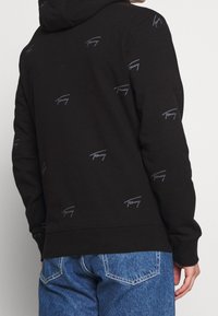 Sudadera negra con un patrón repetido del logo gris "Tommy". Presenta capucha, puños acanalados y un bolsillo tipo canguro. Fabricada en un tejido suave y texturizado.