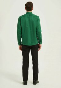 Chemise verte à manches longues, tissu lisse, col standard, poignets à bouton et pli au dos. Associée à un pantalon noir et des chaussures.