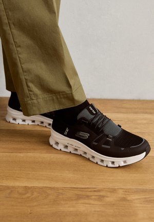 Zwarte sport schoenen met mesh accenten en een witte zool met een uniek loopvlakontwerp. Gecombineerd met kaki broeken.