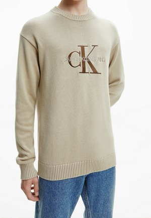Personne portant un pull beige Calvin Klein à manches longues avec un ourlet côtelé, associé à un jean en denim bleu.
