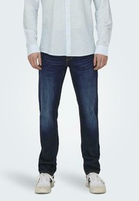 Jeans in denim blu scuro con vestibilità comoda, caratterizzati da un leggero sbiadimento e un classico design a cinque tasche, abbinati a sneaker atletiche bianche.