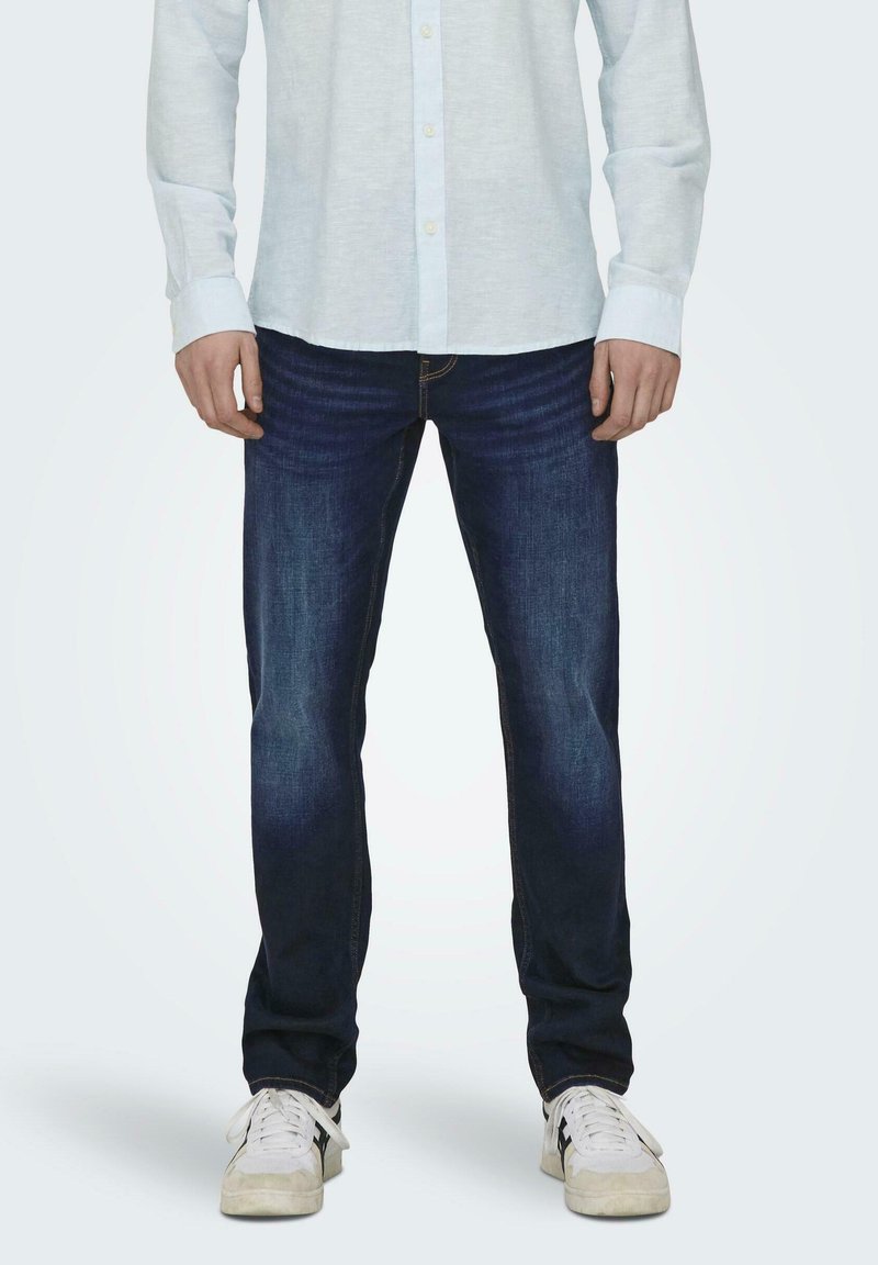 Jeans in denim blu scuro con vestibilità comoda, caratterizzati da un leggero sbiadimento e un classico design a cinque tasche, abbinati a sneaker atletiche bianche.