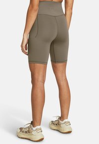 Femme portant des shorts de vélo taille haute taupe et des chaussures de sport beiges, se tenant de dos face au spectateur sur fond blanc.