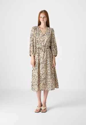 Beige dress with black spotted pattern, V-neck, puff sleeves, and a cinched waist. Worn with light-colored sandals. Smooth texture.
