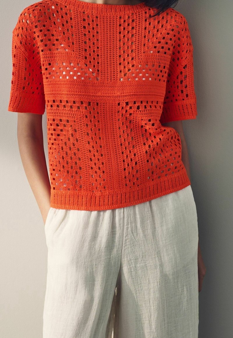 Femme portant un haut crochet à manches courtes orange vif avec des motifs géométriques et un pantalon large en lin blanc, les mains dans les poches, debout contre un mur.
