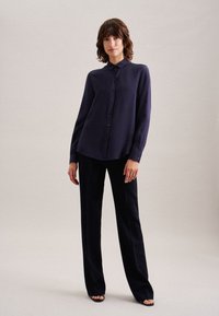 Marineblauwe longsleeve shirt met een knoopsluiting aan de voorkant, gemaakt van een gladde, lichte stof, gecombineerd met zwarte wijde broek en sandaal met open teen.