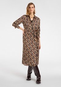 Długa sukienka w kolorze brązowym z wplecionymi geometrycznymi wzorami w kolorze kremowym, pomarańczowym i złotym, z wiązanym paskiem w talii i zapinana na guziki z przodu. Śliskie, sięgające do kolan botki.