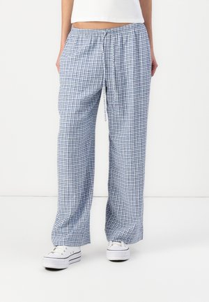 Mujer que lleva pantalones holgados a cuadros azules y blancos con cintura ajustable y zapatillas deportivas blancas con plataforma, de pie frente a un fondo liso.