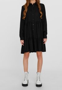 Woman wearing a long-sleeve black buttoned tiered dress paired with white lace-up ankle boots against a plain background.