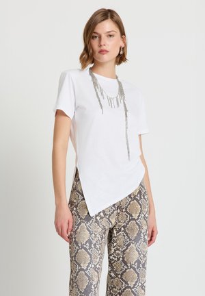 Donna che indossa una T-shirt bianca asimmetrica, pantaloni con stampa a serpente, collana con frange d'argento e orecchini a cerchio d'argento, in piedi davanti a uno sfondo semplice.