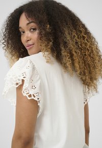 Haut en coton blanc avec des manches volantées bordées de dentelle. Texture douce avec une coupe décontractée, mettant en valeur des détails perforés complexes sur la dentelle.