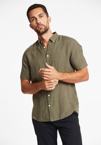 Olijfgroene kortemouwige blouse met knopen van linnenstof, voorzien van een puntige kraag en een subtiele textuur, gestoken in donkere broeken.
