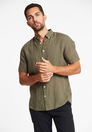 Olijfgroene kortemouwige blouse met knopen van linnenstof, voorzien van een puntige kraag en een subtiele textuur, gestoken in donkere broeken.